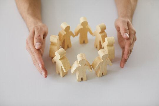 Human Hands Protecting And Guarding Small Wooden Toy Figures Placed On White Desk As Metaphor For Creating Safe, Supportive Community Of People. Close Up, Closeup Shot. Care, Support, Safety Concepts