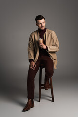 Full length of stylish man in autumn outfit holding paper cup while sitting on chair on grey background.