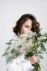 A mysterious girl with wavy hair in a white blouse covers her face with a bouquet of flowers on a...