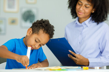  Little boy, training in kindergarten, sitting at the table with a friendly teacher and doing...