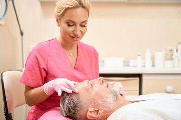 Fototapeta premium Smiling woman cosmetologist doing facial massage of man