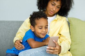 Little ethnic boy watching cartoons on phone smartphone with mother while sitting on couch at home...