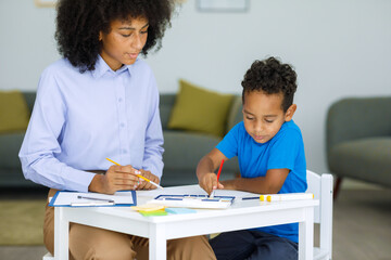 African american little boy painting in kindergarten, sitting at a table with a teacher and holding a paintbrush, a woman is doing arts and crafts with a baby in kindergarten.
