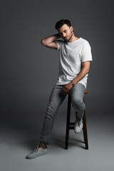 Full length of young man in t-shirt and jeans sitting on chair on grey background.