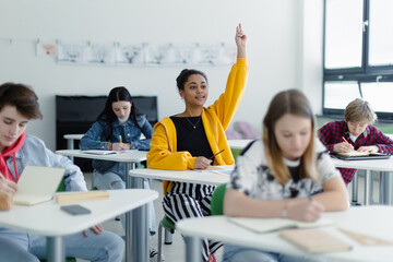 High school students paying attention in class, sitting in their desks and raising hand.