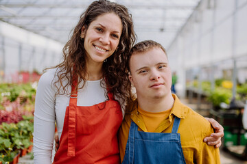 Experienced woman florist posing with young colleague with Down syndrome in garden centre.