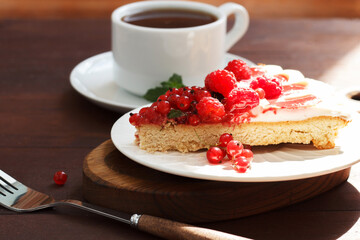 A piece of berry pie with cheese cream on a plate on a wooden background with a cup of tea.