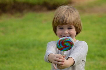 Emotions of a child. Portrait of a little positive boy with a big candy on a stick. Multi-colored lolipop in the hands of a child. Summer day, sincere joy, holiday, day off, walk with parents, grandmo