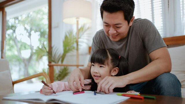 
Asian young kid daughter coloring and painting on paper with father. Happy family activity, Little girl children learn to draw art picture enjoy creativity in weekend with dad in living room at home.