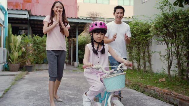 Asian attractive couple parent teaching kid daughter riding on bike. Young father and mother giving teach little girl child practice and learning ride bicycle with safety helmet in backyard at home.
