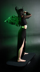 portrait of beautiful asian model with dark hair, wearing black gothic skirt costume, angel feather wings with horned headdress. Posing with gestural hands  on dark silhouette  studio background.