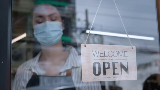 Caucasian beautiful waitress wear mask, turns open sign on glass door. Attractive Coffee shop owner prepare to welcome customer to reopen small business after COVID-19 ended lockdown and quarantine.
