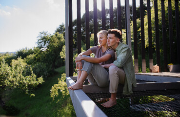 Young couple sitting and cuddling in hammock terrace in their new home in tiny house in woods,...
