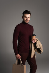 Stylish brunette man with jacket holding coffee to go and shopping bag isolated on grey.