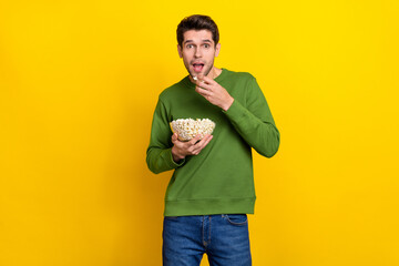Photo of positive impressed young guy wear green sweater eating tasty popcorn isolated yellow color background
