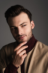 Brunette man in jacket touching chin and looking away isolated on grey.