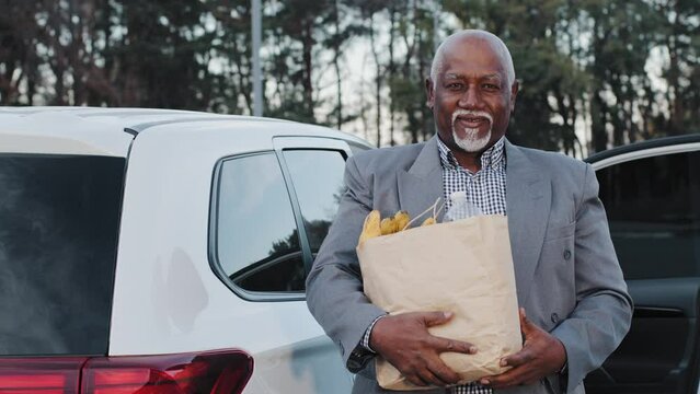 Handsome old african american man smiling holding food in hands bought in fruit shop happy carefree pensioner male closeup stands in front white car elderly person looking at camera gray hair beard