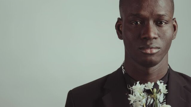 Studio portrait of stylish African American man wearing black jacket and floral pattern shirt holding fresh white flowers looking at camera