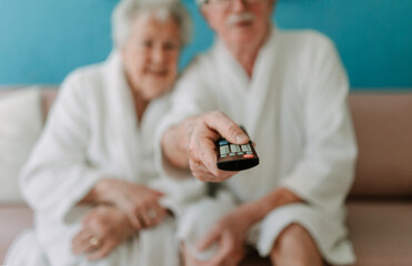 Happy senior couple sitting at sofa in bathrobes and watching TV.