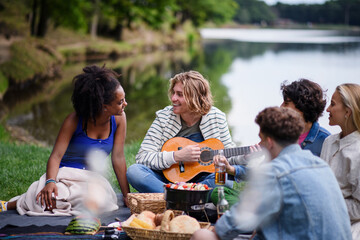 Group of friends having fun on picnic near a lake, sitting on blanket eating and playing guitar.
