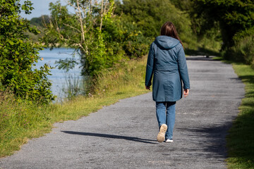 Woman walking on a footpath in a park by a river. Back to camera. Warm sunny day. Outdoor activity.