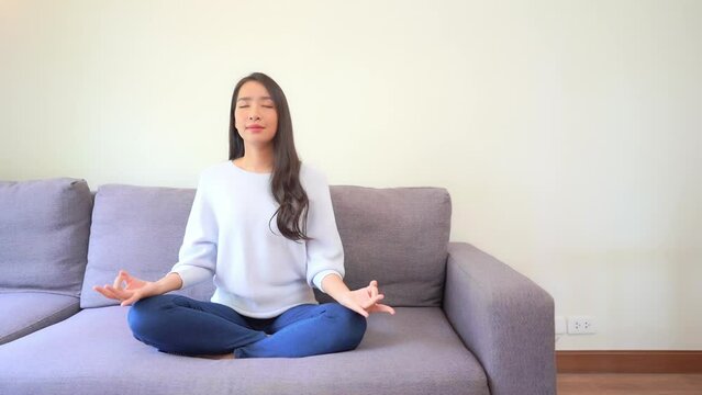 Dressed in jeans and a sweater, a young woman with closed eyes strikes a yoga pose on a couch. Title space
