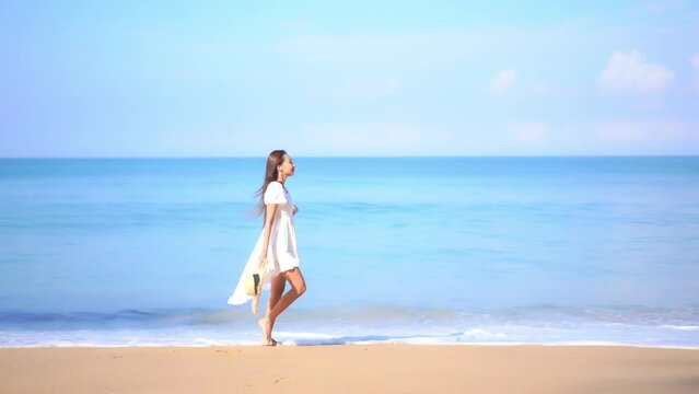 Attractive Woman in White Dress With Hat in Hand Walking on Sandy Beach in Front of Tropical Sea, Slow Motion