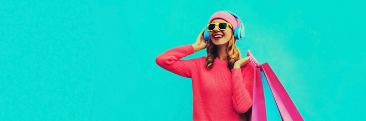 Portrait of stylish happy smiling young woman enjoying listening to music in headphones with colorful shopping bags posing wearing pink knitted sweater, hat on blue background