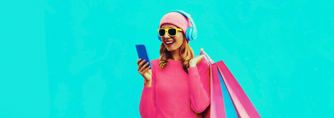 Farbenfrohes Porträt einer stilvoll lächelnden jungen Frau, die Musik über Kopfhörer mit Smartphone und Einkaufstaschen hört, die Strickpullover tragen, rosa Hut auf blauem Hintergrund