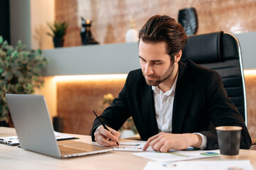 Concentrated clever caucasian business man, IT specialist, ceo, sits at the workplace in the office, works in a laptop, works on a new project, develop new app, strategy, study documents, graphs