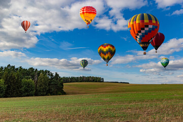 Multicolored hot air balloons fly in blue sky with white clouds over green field.
