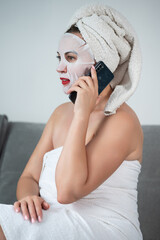 A woman is talking on the phone at home, a woman with a mask on her face and a towel is sitting on the sofa.