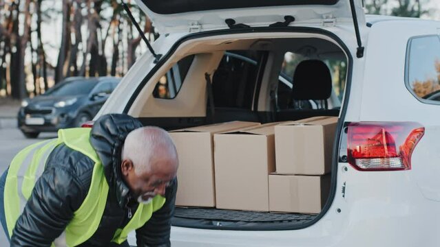 Close-up mature delivery service worker unloading cardboard boxes from trunk of car elderly african american courier loader delivering goods handyman smiling in work clothes accepts shipping outdoors