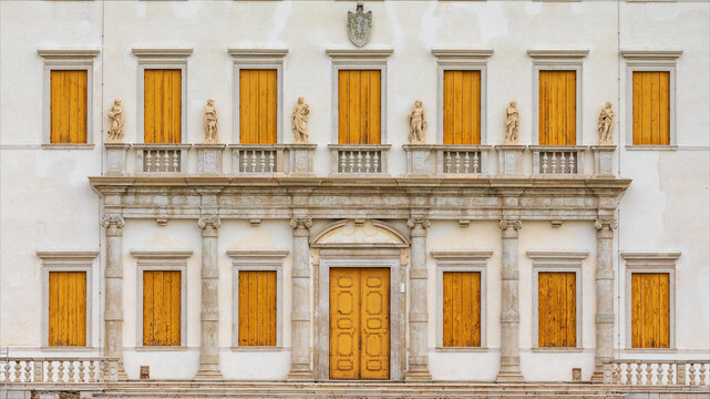 Numerose finestre di legno chiuse, statue e una porta su facciata di antico edificio storico in stile veneziano di colore bianco.
