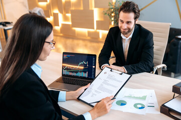 Cooperation concept. Hiring specialist in a trading company, sits at a desk in front of a laptop, conducts a hiring interview with clever caucasian man, focused studying candidate's resume