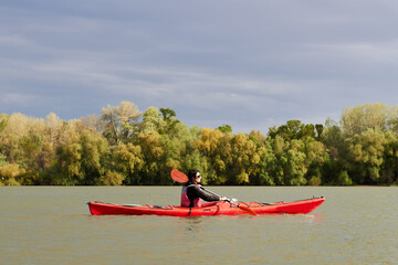 A girl on a red kayak paddles along the Danube river in spring against dark blue sky and gently green trees. Ukraine