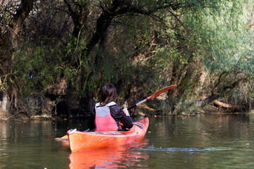 Rear view on woman paddle red kayak in spring river against gently green willow trees