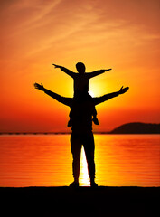 silhouette of a father with a child on his shoulders, arms outstretched at sunset