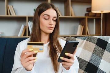 Happy young woman holding phone and credit card sitting on the couch. Online shopping.