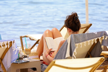Woman in bikini reading book sitting in deck chair on a sea beach. Summer relax on luxury resort