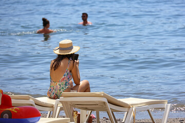 Girl in swimsuit ad sun hat sitting with smartphone in hands in deck chair on sea waves and...