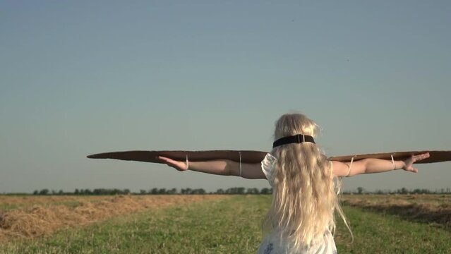 Girl child in airplane pilot glasses with cardboard wings runs through a rural field, slow motion