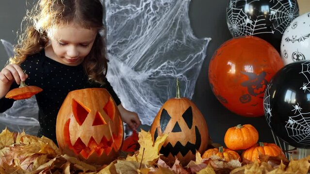A little curly girl of European appearance pulls candy out of a halloween pumpkin with a funny face on a black background with spider webs, autumn leaves and balloons. Trick or treat