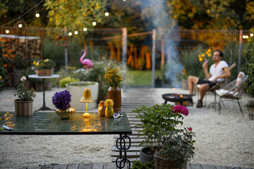 Beautiful and cozy garden with man relaxing sitting by the fireplace on background at dusk