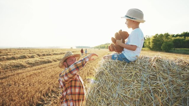 Man stands in wheat field and points finger to beautiful summer nature in wheat field. Boy sits on haystack and looks into distance, holding toy teddy bear in hands. Leisure activity parent and child.