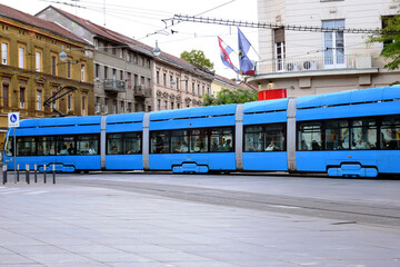 Naklejka premium long blue tram in side perspective view in Zagreb, Croatia. popular square in downtown. old classic residential buildings and stucco facades in the background. travel and tourism concept. urban scene.