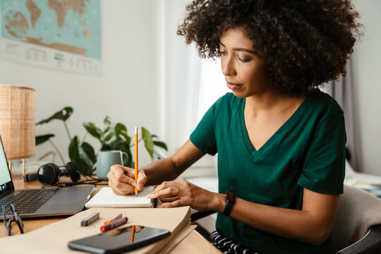 African American Young Woman Using Laptop And Taking Notes At Home