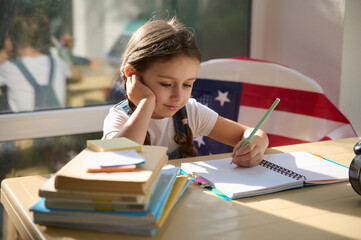 Charming little girl with two pigtails, cutely smiles, enjoys learning on first grade, doing homework, sitting on a chair with an American flag, at a desk with textbooks and stationery on the table