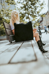 Blonde women sitting on bench at the city with black textile handbag, recycled textile handbag