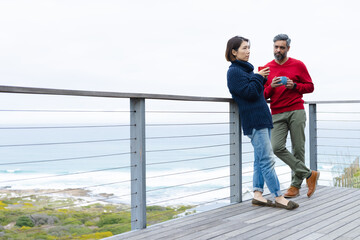 Diverse couple holding mugs of coffee and wearing jumpers together at balcony